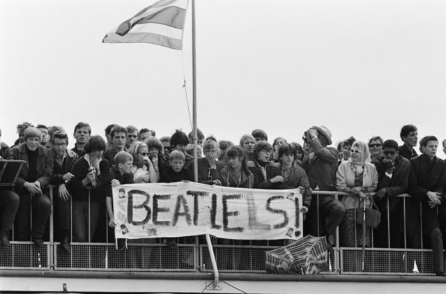 Nuoret fanit odottavat idoleitaan The Beatlesin saapuessa Schipholiin. Bannerin tekijät ovat luultavasti yrittäneet korjata alkuperäistä kirjoitusvirhettä. Photo Credit