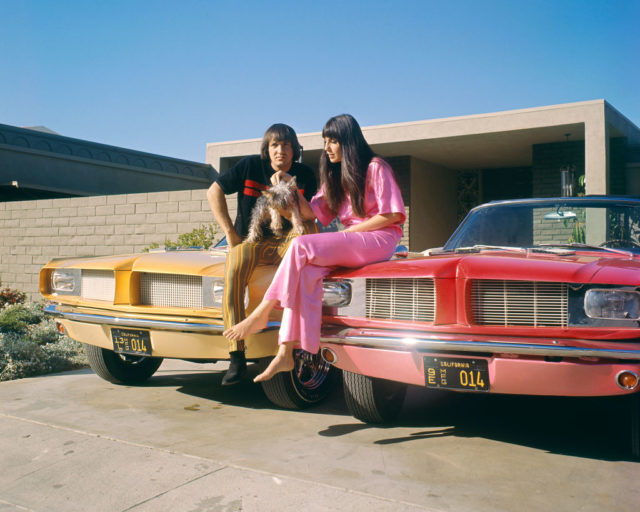 Sonny Bono and Cher sitting on two cars with a dog