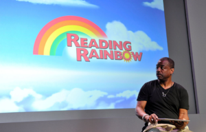 LeVar Burton sitting in front of the Reading Rainbow logo