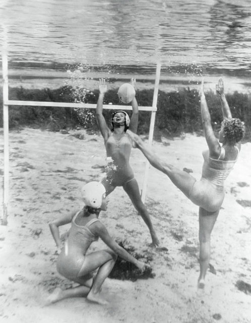 Black and white photo of three women in bathing suits playing vollyball under water.