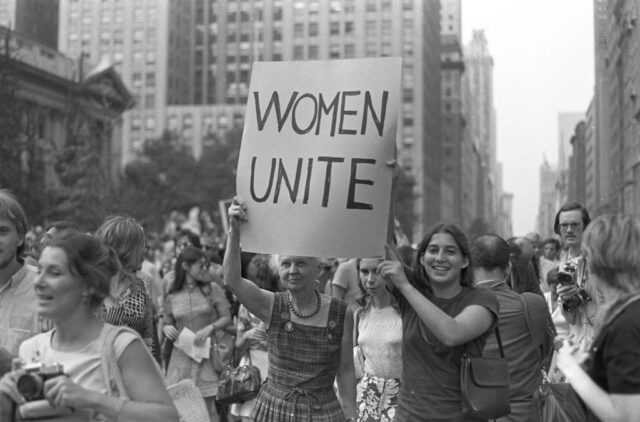 Women marching down a street, with one holding up a sign that reads, "WOMEN UNITE"