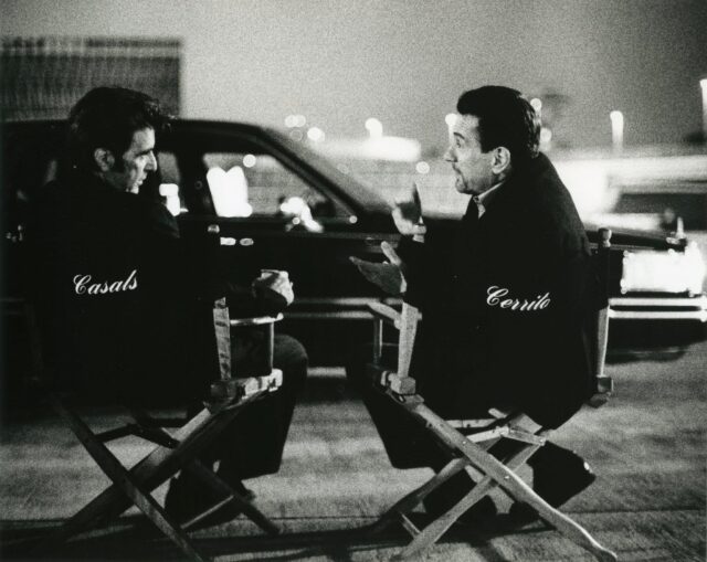 Al Pacino and Robert DeNiro sit on directors chairs talking to each other with a car in the background.