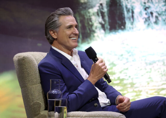 Gavin Newsom sitting in a chair on stage, holding a microphone