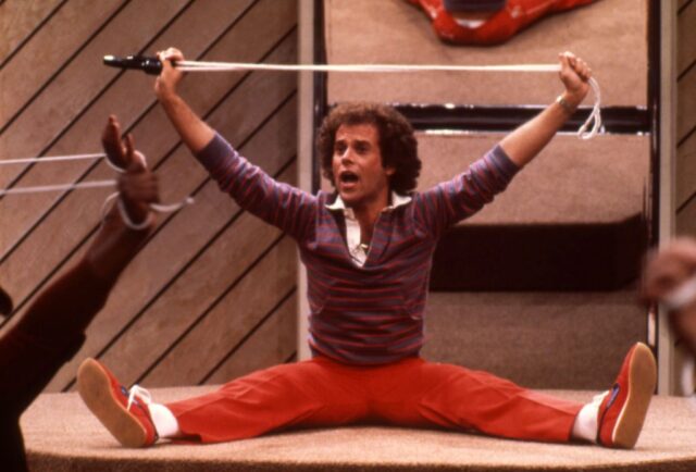 Richard Simmons sitting on a gym mat, holding a rope over his head