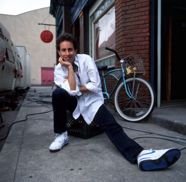 Jerry Seinfeld sitting with his leg out.