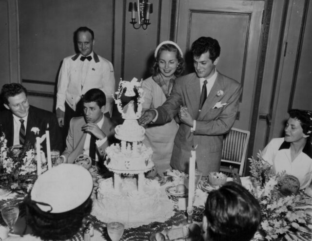 Janet Leigh and Tony Curtis cut the cake during their wedding, people sitting around them.
