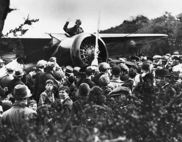 Amelia Earhart waves at a crowd surrounding her plane.