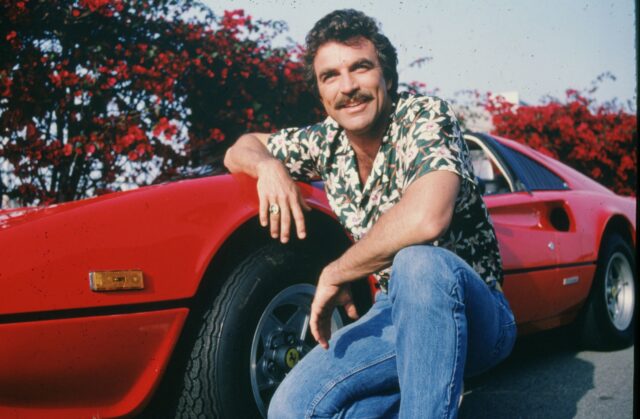 Tom Selleck crouching by a red car.
