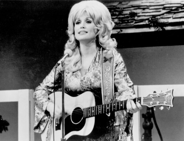 Dolly Parton at a microphone playing the guitar.