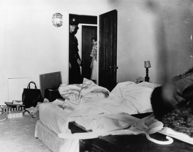 A messy bedroom, two people standing in its doorway looking in.