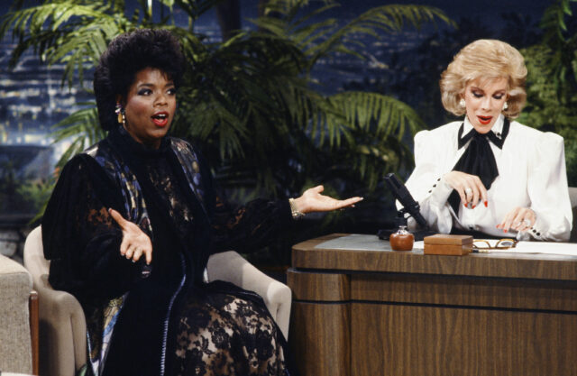 Oprah Winfrey sitting beside Joan Rivers sitting behind a desk.