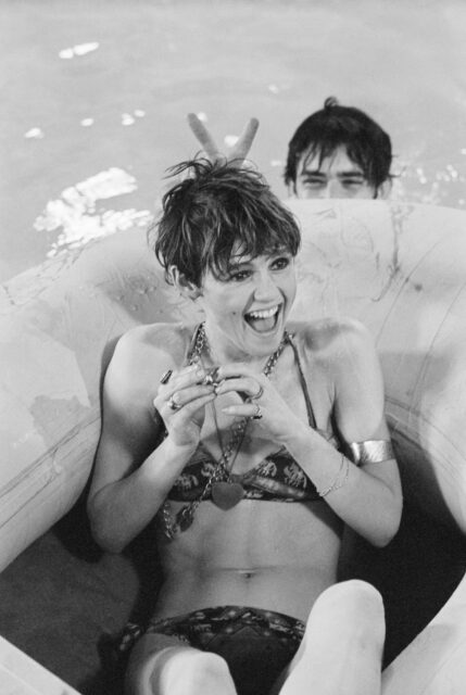 Edie Sedgwick on a pool float while a man holds up his fingers behind her head to look like bunny ears