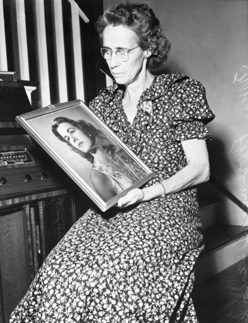 Florence Spangler sitting near a staircase, holding a portrait of Jean Spangler