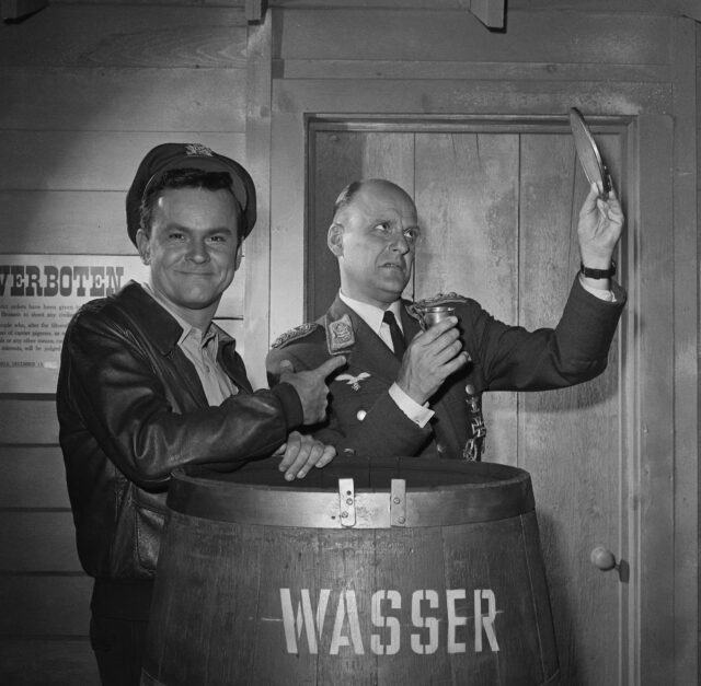 Bob Crane and Werner Klemperer behind a wood barrel in army costume.