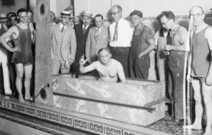 Harry Houdini waves at the camera while sitting in a coffin, people surrounding him