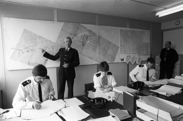 Inspector Peter Smith pointing at a map, while other police officers work at a desk in the middle of a room