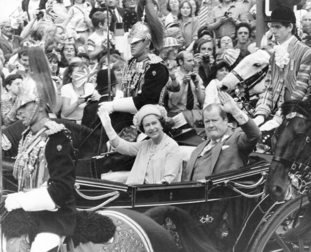 Queen Elizabeth II and Edward John, 8th Earl Spencer waving to a crowd of people standing around the horse-drawn carriage they're riding in