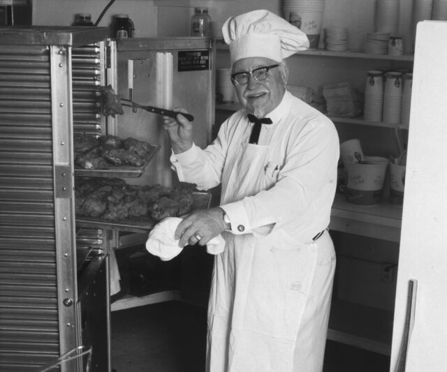 Colonel Sanders of KFN holding fried chicken in a kitchen.