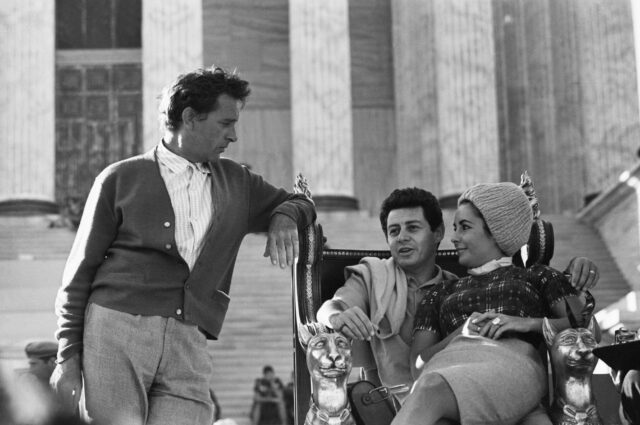 Elizabeth Taylor sitting on Eddie Fisher's lap, while Richard Burton stands beside them