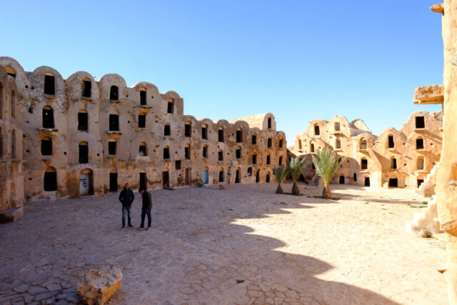 Two people stand in the courtyard of tall hut buildings with many stories.