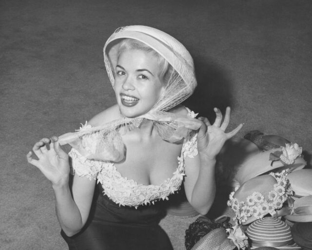 Jayne Mansfield kneeling next to a pile of bonnets, while wearing one on her head