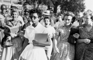 An African American woman walks with books in her hand as a crowd gathers around her screaming.