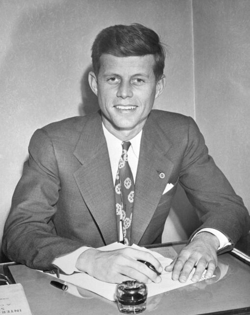 Headshot of John F. Kennedy sitting at a desk with pen and paper in hand.