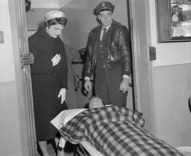 Jackie Kennedy and a police officer standing over JFK laying on a gurney.