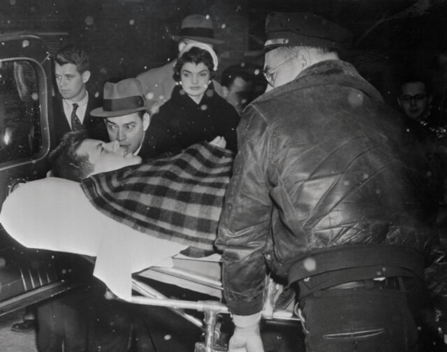 JFK laying on a gurney, Jackie Kennedy and other surrounding him.