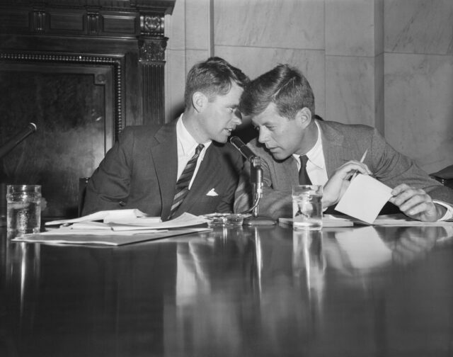 Robert Kennedy whispering something into John F. Kennedy's ear at a table. 