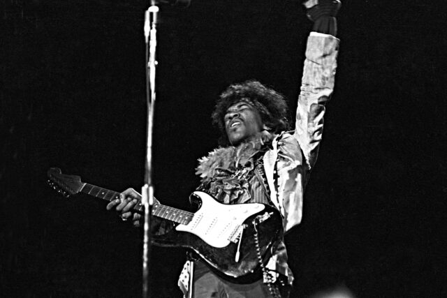 Jimi Hendrix with his arm up, a guitar slung from his shoulder.