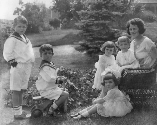 Some of the Kennedy children with their mother, John F. Kennedy sitting on a wooden bike.