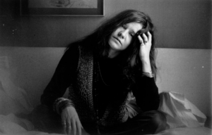 Janis Joplin sitting cross-legged with her head in her hand.