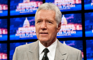 Alex Trebek standing in front of several screens reading 'Jeopardy.'