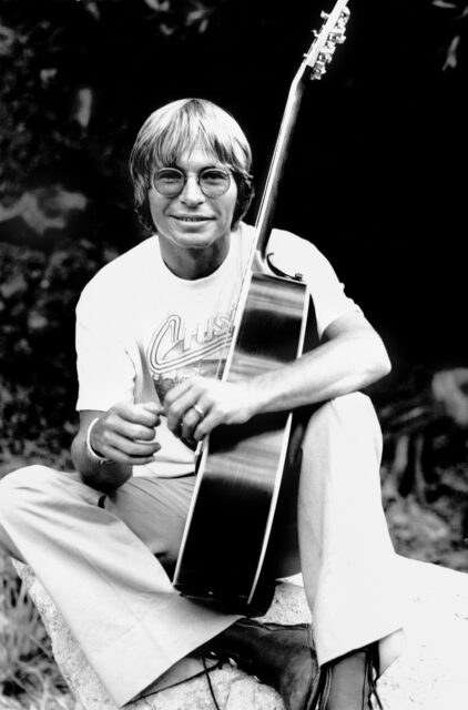 John Denver sitting with his guitar.