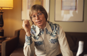 John Denver sitting in a room with his one hand raised near his face.