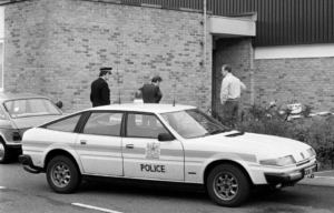 A police car parked in front of a brick building, three officers standing around it.