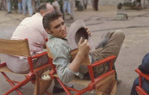 Elvis sitting in a chair on set of a film, holding a hat.