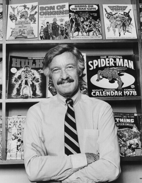 A young Stan Lee stands in front of a shelf displaying several comic books.