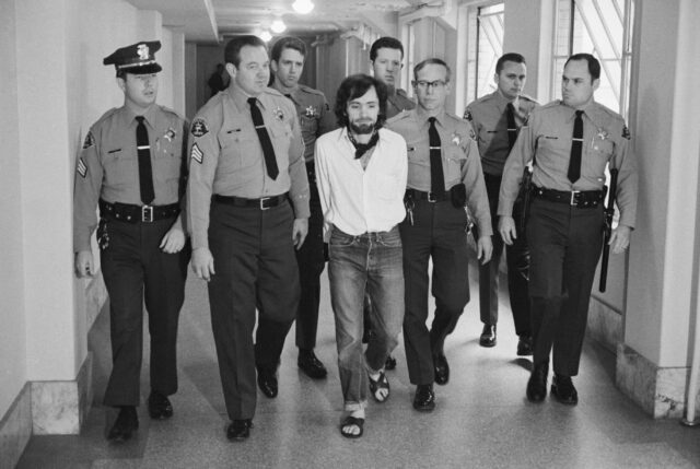 Charles Manson being escorted in a hallway by several officers.