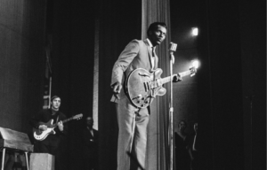 Chuck Berry performing into a microphone with a guitar.