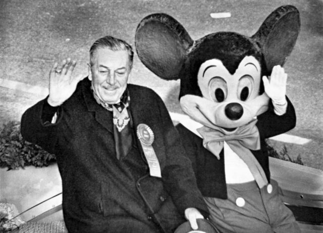 Walt Disney sitting beside a Mickey Mouse mascot, both waving.