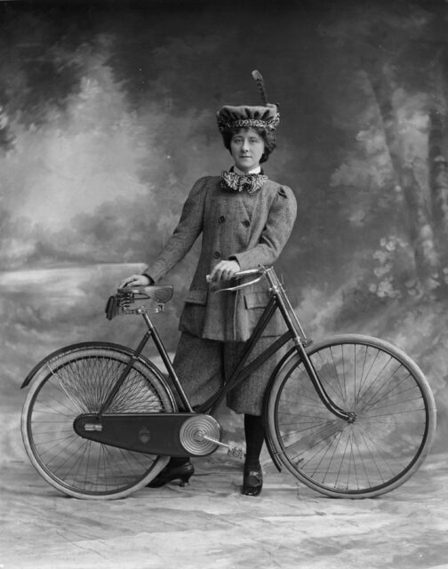 A Victorian woman stands with her bicycle, wearing pants.