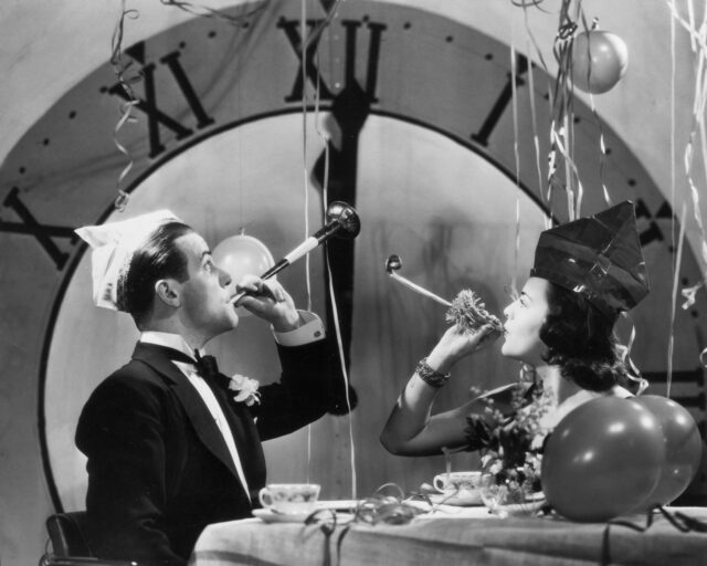 A man and woman blowing horns wearing celebration hats in front of a clock.