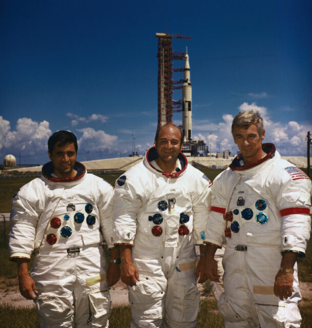 Three male astronauts in front of a rocket ship.